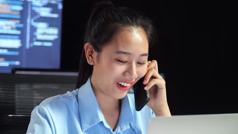 close up of asian female programmer talking on smartphone while writing code by a laptop using multiple monitors showing database on terminal window desktops in the office