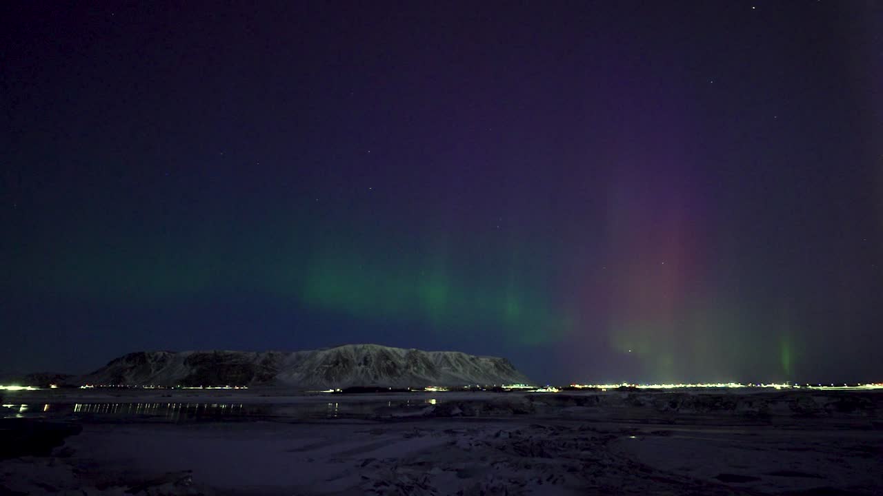 Time lapse shot of flickering green and purple aurora borealis at night sky of Iceland. Traffic on snowy winter road. Wide shot. Lighting city at horizon.