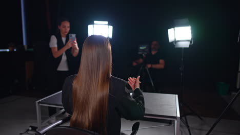 rear view of an unrecognized woman with long brunette hair sitting at desk in the light of soffits. cameraman behind the professional camera and girl with a phone filming at backdrop.