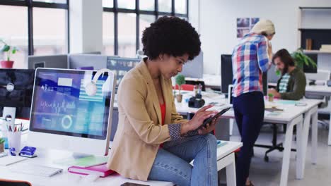 Mixed-race-businesswoman-sitting-on-desk-using-digital-tablet-in-office