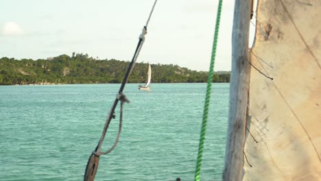 landscape shot from a rustic sail boat with detail of ropes and dirtied by time sails, in a bright blue cozy water area, with natural green forest land and sail boat in the horizon on a sunny day