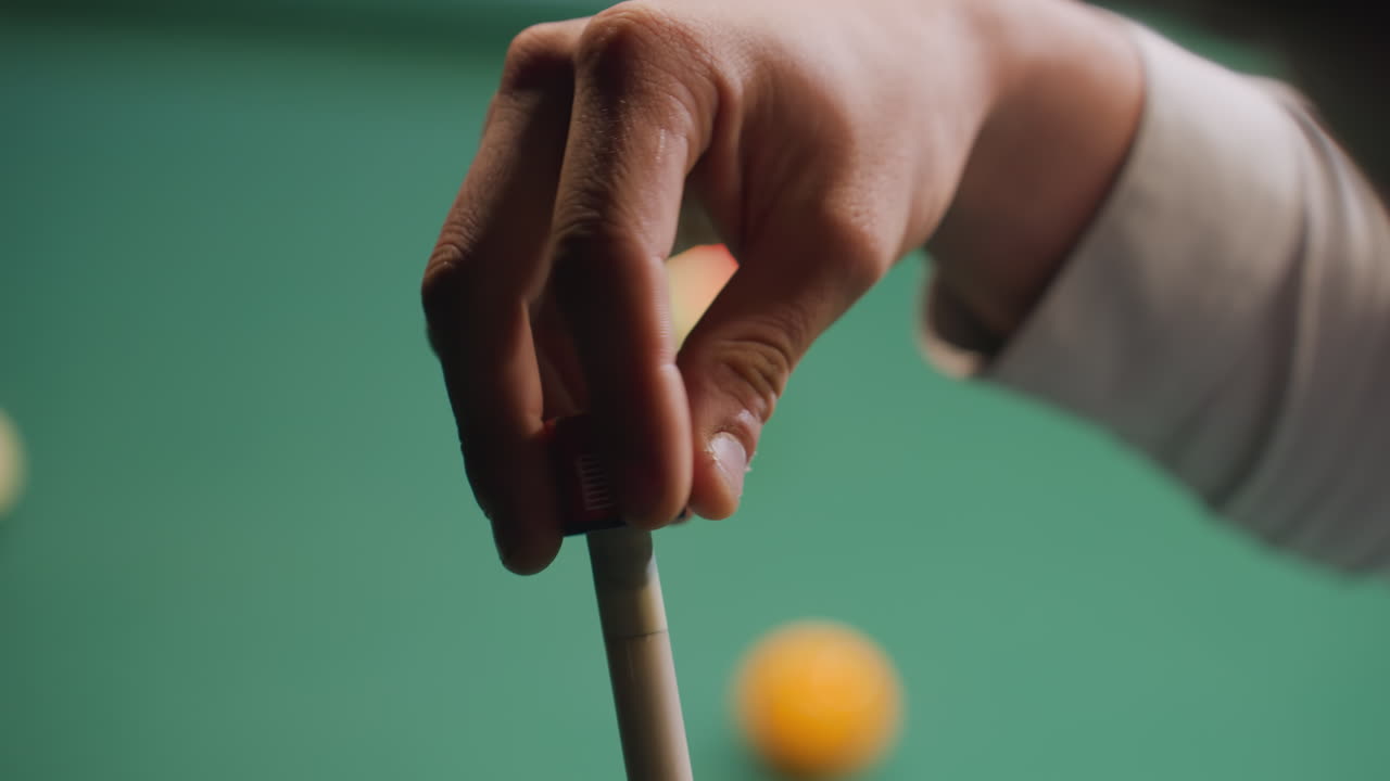 Close-up da mão de um homem passando giz em um taco de sinuca antes de uma tacada em uma mesa de bilhar verde. Foco na precisão e na técnica, com bolas de bilhar borradas ao fundo. Movimento suave, jogo profissional