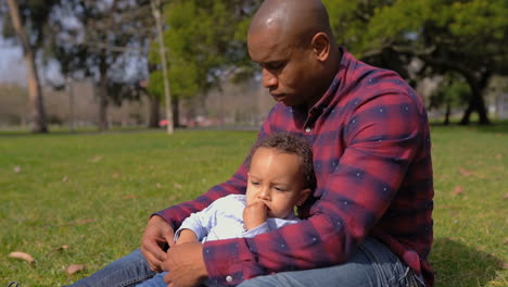 father sitting on grass in park, holding son on knees, hugging