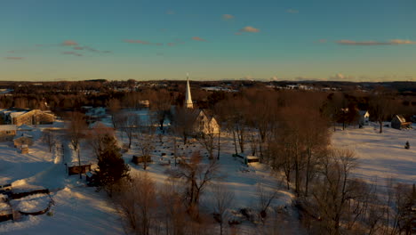 Scenic-winter-aerial-flying-over-a-picturesque-snow-covered-small-town-at-sunset