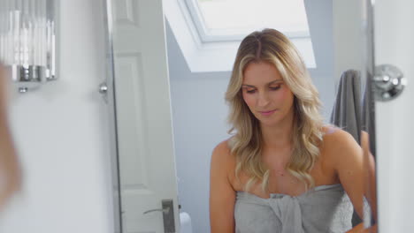 mature woman putting on moisturizer looking at reflection in bathroom mirror at home wearing towel