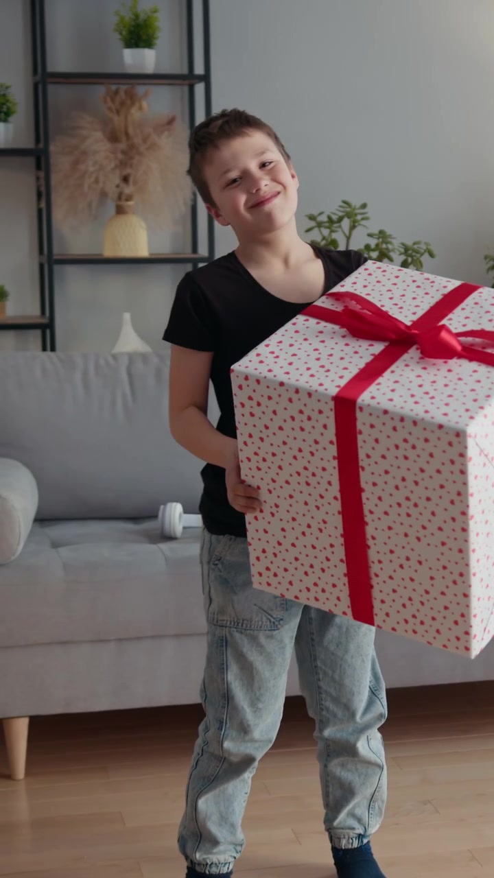 Un ragazzo in abiti casual porta una scatola regalo molto grande e decorata con un nastro rosso attraverso un soggiorno.