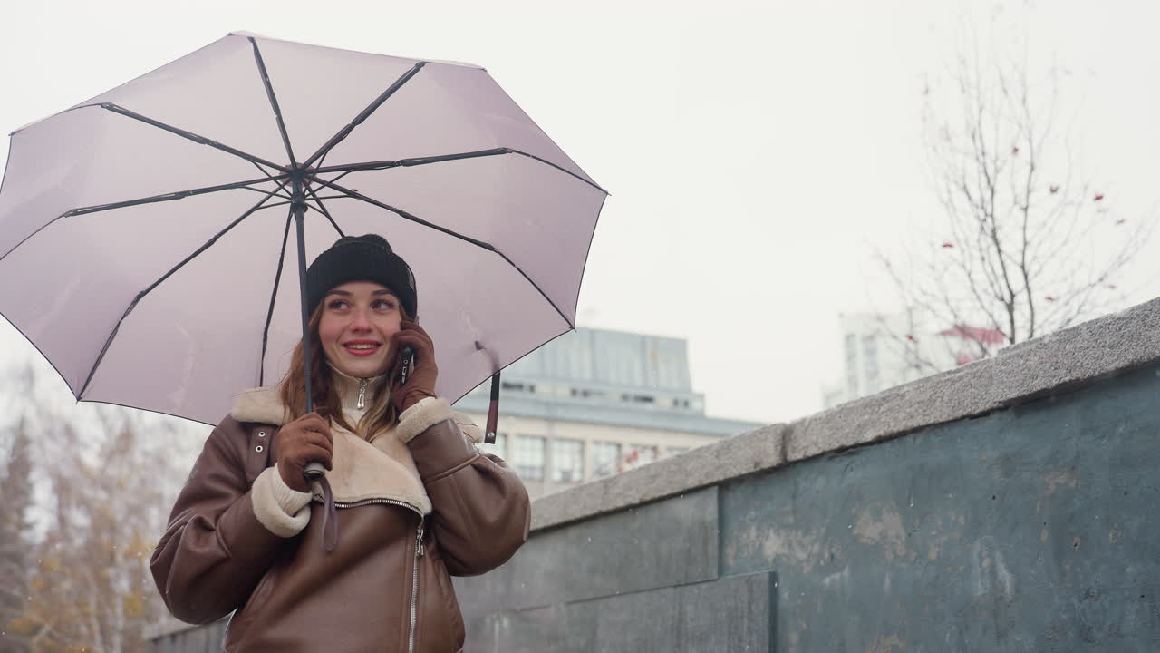 Cheerful girl smiling on phone call holding umbrella, wearing knit cap, brown shearling jacket, walking in light snowfall with soft background of autumn trees and urban setting