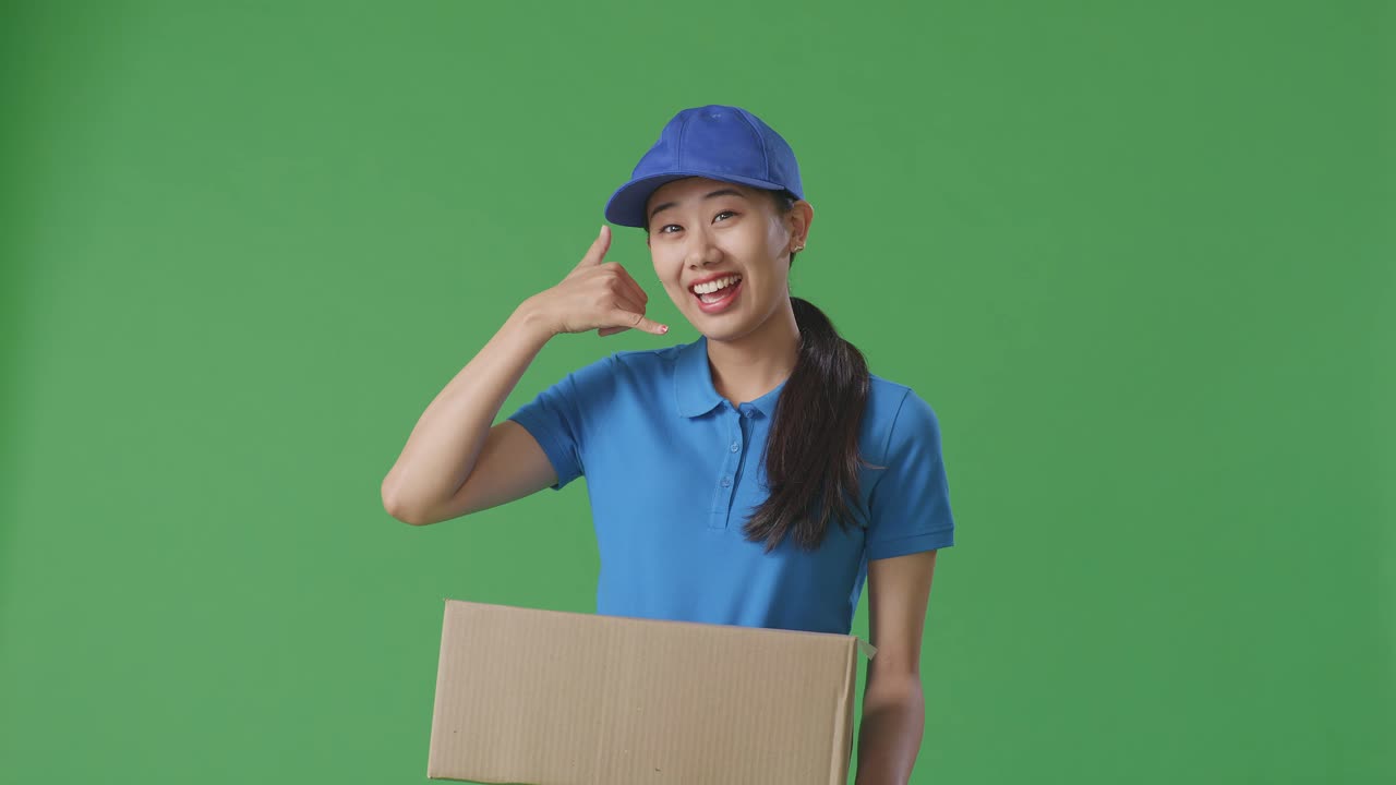 Asian Female Courier In Blue Uniform Showing Call Me Gesture And Smiling While Delivering A Carton On Green Screen Background In The Studio