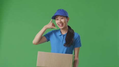 푸른 유니폼을 입은 아시아 여성 택배기사는 스튜디오에서 녹색 화면 배경에 상자를 배달하는 동안 제스처와 미소를 지으며 나를 호출합니다.