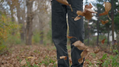 vista de ángulo bajo de una persona con pantalones de jean de pie en el bosque mientras vierte hojas secas de una bolsa en el suelo, con follaje de otoño esparcido en el fondo durante la temporada de otoño