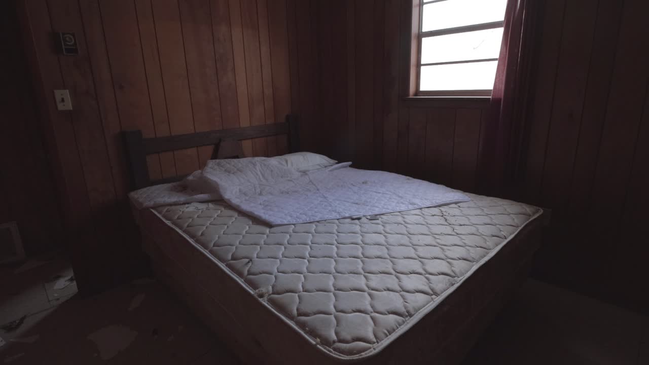 An Unused Bed in an Abandoned Cabin.  Static Shot Showing the Bed, Wooden Walls, and the Broken Floor in the Background.