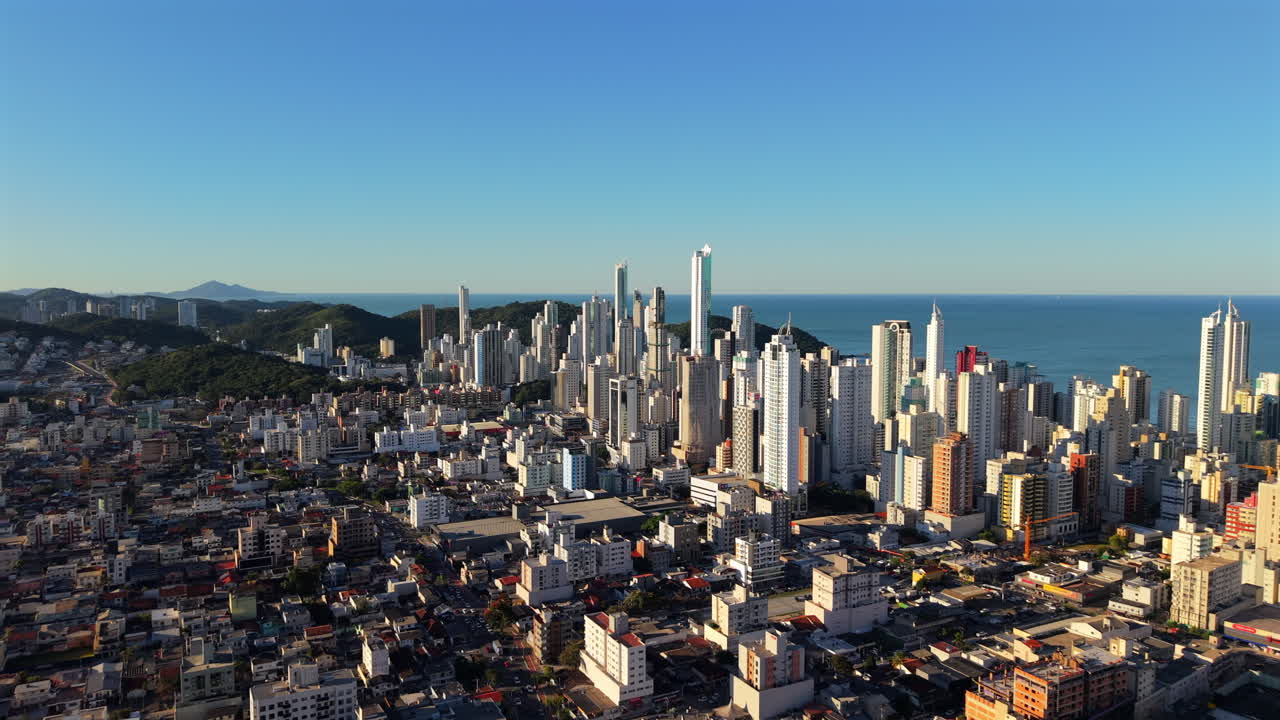 High-altitude daisy cutter drone shot glides toward Camboriú’s sunlit skyscrapers, with their facades shimmering under crisp morning light and ocean background. Ideal for city branding