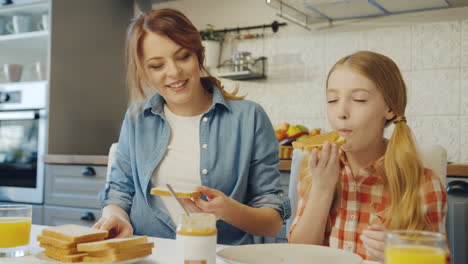 Porträtaufnahme-Der-Hübschen-Mutter,-Die-Sandwiches-Mit-Erdnussbutter-Zubereitet,-Und-Ihrer-Lieben-Tochter,-Die-Sie-Mit-Einem-Lächeln-Isst.-Drinnen