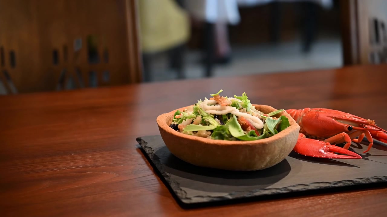 Fresh leafy salad in small brown bowl sits on dark slate beside bright red crab on polished wooden table, with softly blurred restaurant chairs and warm ambient light creating upscale dining mood