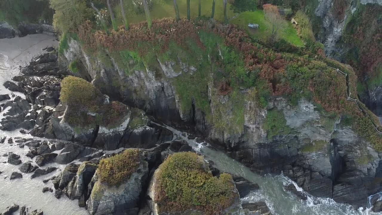 inclinación aérea de drones desde las olas rompiendo en la formación rocosa hasta una casa en un bosque sentado en la cima de un acantilado - lee bay, beach, ilfracombe, devon, inglaterra
