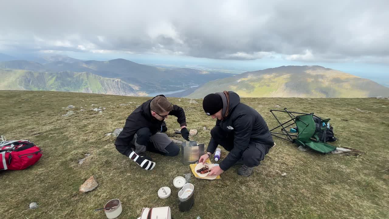 Two campers set up stove and cook near Y Garn summit in Snowdonia with wide mountain view, lake below, and cloudy sky, capturing peaceful outdoor moment in high remote natural landscape
