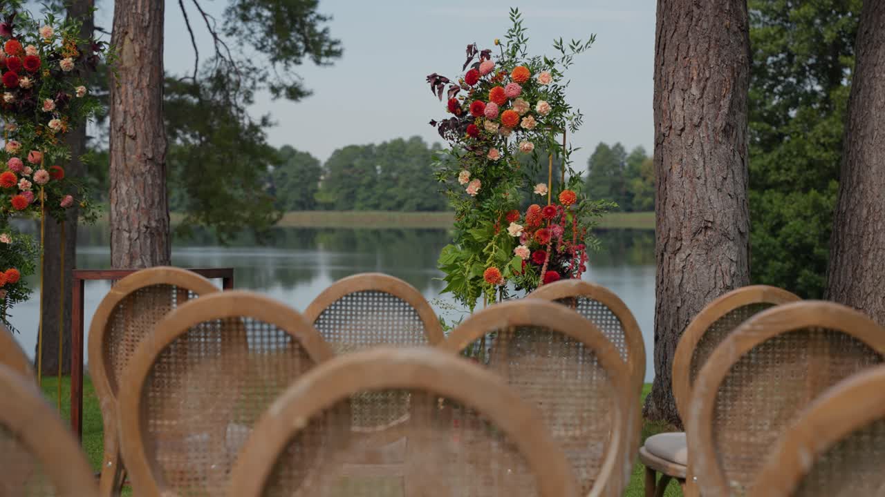 A beautifully decorated outdoor wedding ceremony setup by a serene lakeside, featuring elegant floral arrangements. The lush greenery and calm water create a romantic atmosphere