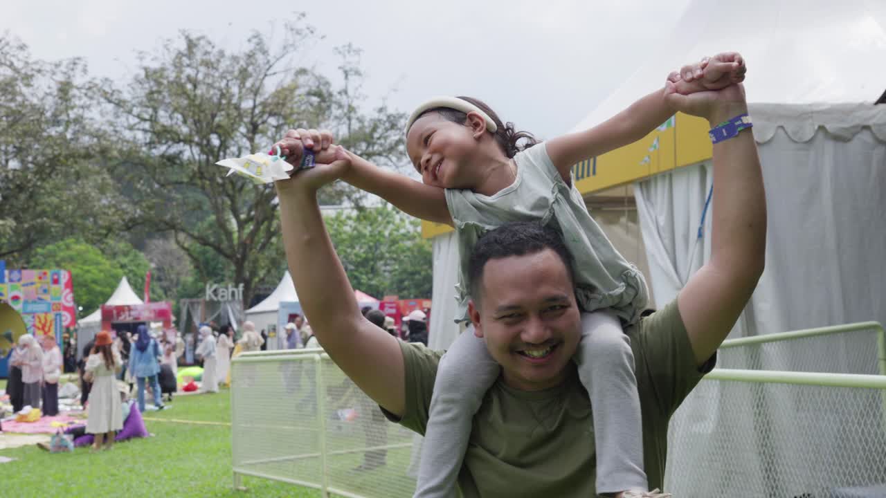 Little girl, perched on her father's shoulders, watches sunset at Bogor Botanical Gardens concert in Indonesia