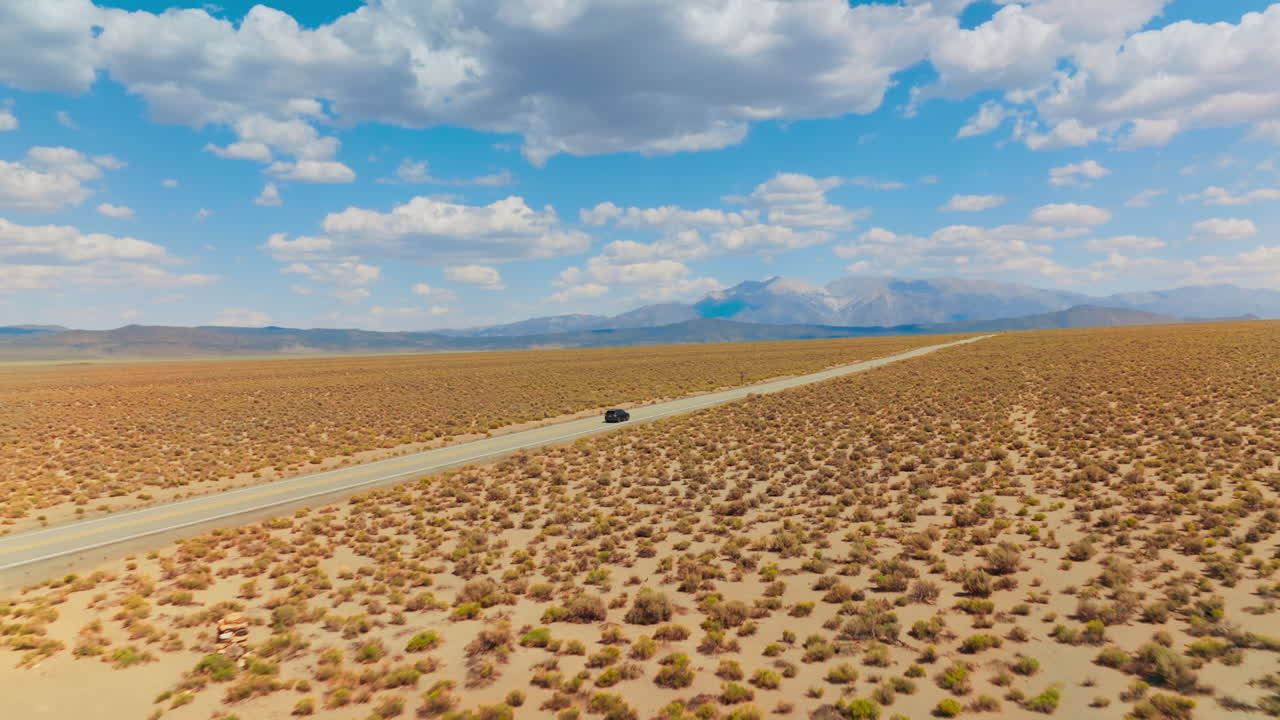 Yellow dry deserted landscape contrasting with bright azure sky with multiple clouds. Black car riding by the road. Mountains at backdrop.
