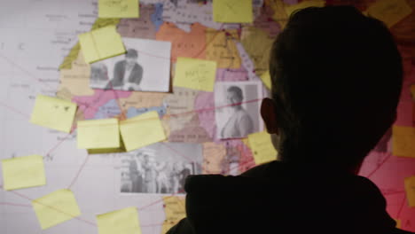 caucasian man looking at wall map with strings, stickers and photos, searching connections