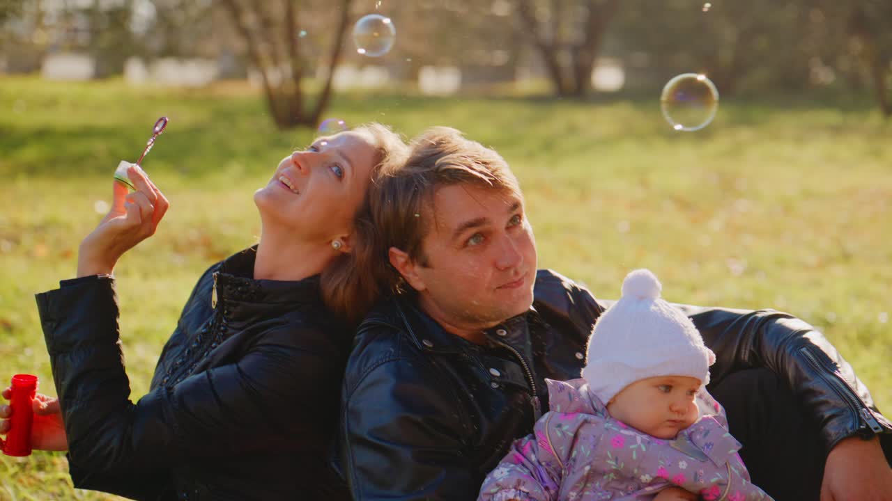 Momenti gioiosi in famiglia nella natura: una coppia felice fa le bolle di sapone mentre è seduta insieme al loro adorabile bambino in una calda e soleggiata giornata in un sereno ambiente esterno