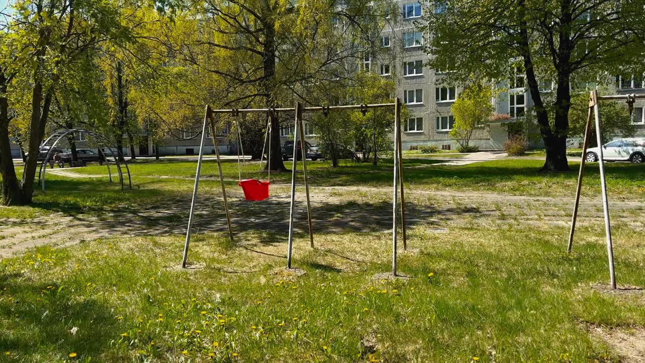 An abandoned red children's swing swings with the wind in summer in a soviet courtyard