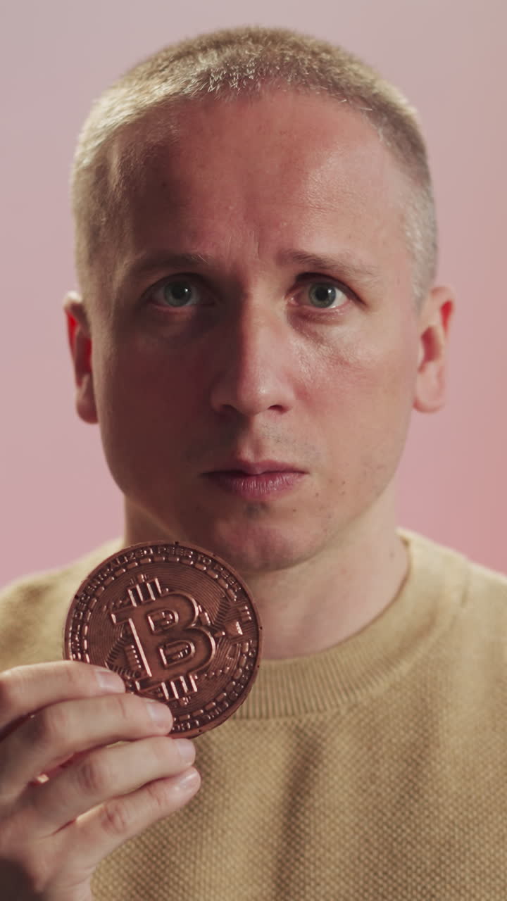 Serious young man holds bitcoin model in hand looking in camera. Portrait of person dealing with cryptocurrency on blurred pink background closeup