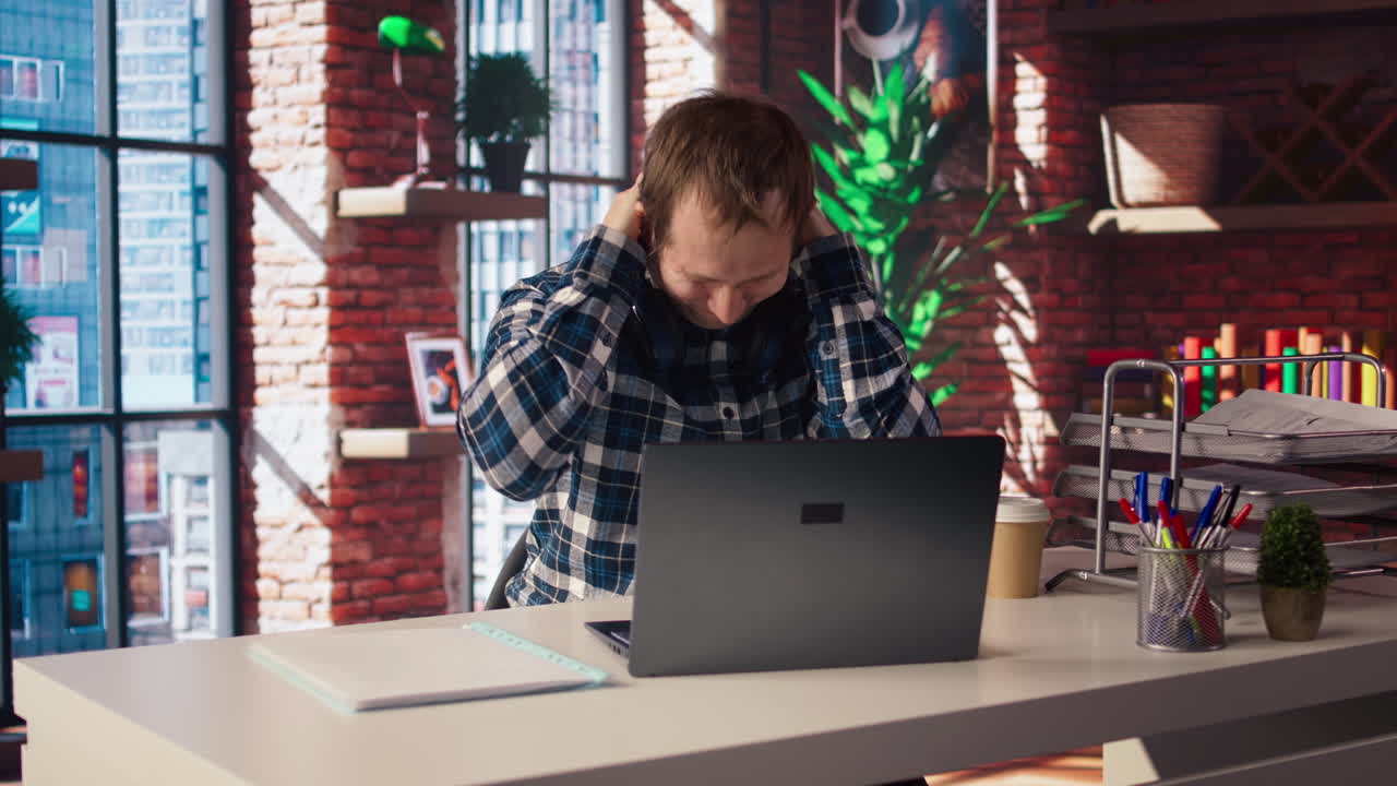 Stressed man in home office scratching head with pen, feeling overworked