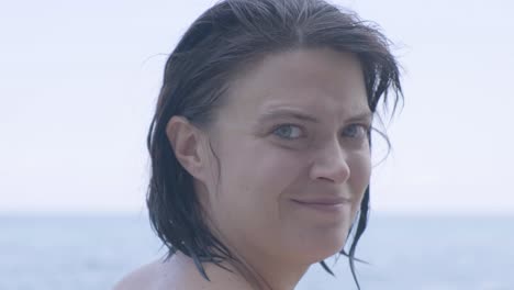Caucasian-Woman-Staring-And-Smiling-At-The-Camera-Brushing-Her-Wet-Hair-Isolated-In-Blurry-Sea-Background---Closeup-Shot