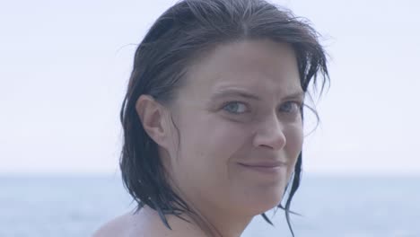 caucasian woman staring and smiling at the camera brushing her wet hair isolated in blurry sea background - closeup shot