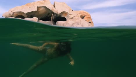 Half-underwater-scene-of-little-girl-with-swimsuit-and-diving-mask-swimming-in-sea-water-of-Cala-Della-Chiesa-lagoon-with-granitic-rocks-in-background-on-Lavezzi-island-in-Corsica,-France