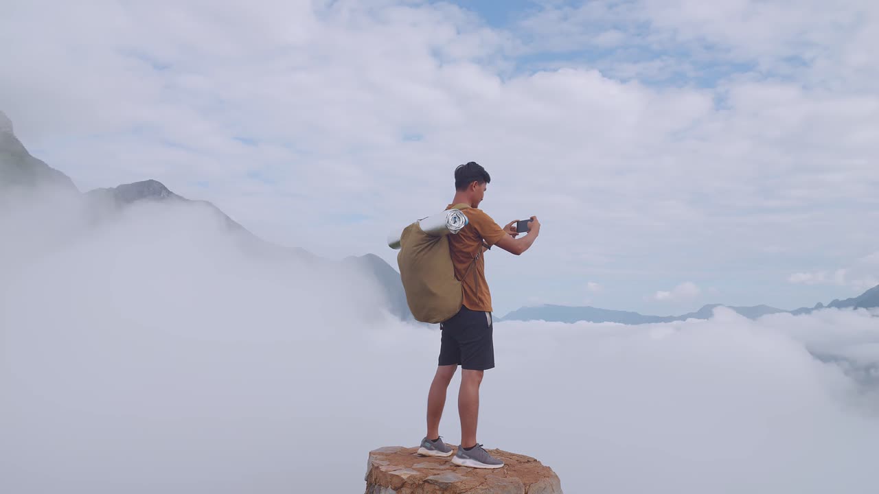 스마트워치를 이용해 산 위에 서서 안개가 가득한 산의 사진을 찍는 아시아인 하이커