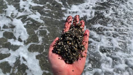 Hand-holding-a-beautiful-pile-of-tiny-little-rocks-at-the-beach,-sea-water-and-waves-in-Manilva-Spain,-fun-sunny-summer-day,-4K-static-shot