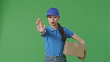 asian female courier in blue uniform making stop gesture and shaking her head while delivering a carton on green screen background in the studio