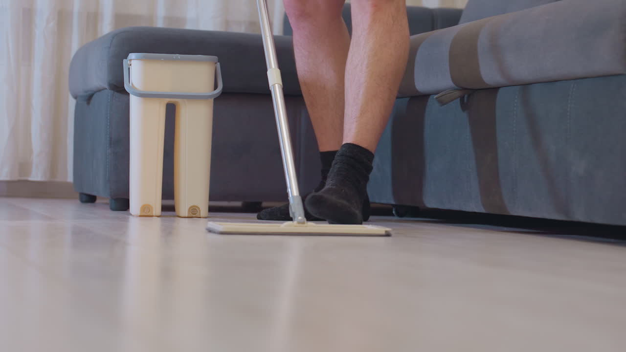 Lower angle view of adult wearing black socks standing on floor holding mop handle while cleaning near gray sofa and bucket in bright room showing household cleaning task