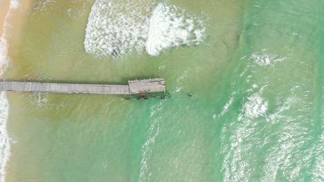 Filmische-Luftdrohnenaufnahme-Von-Klaren-Meereswellen,-Die-Auf-Einen-Leeren-Strand-Mit-Einem-Hölzernen,-Von-Hand-Gebauten-Pier-Als-Zentralem-Thema-Auf-Einer-Asiatischen-Insel-Krachen