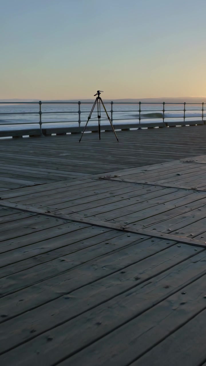 Vídeo vertical: Câmera panorâmica passando por tripé estacionário no píer ao nascer do sol, capturando o brilho do horizonte
