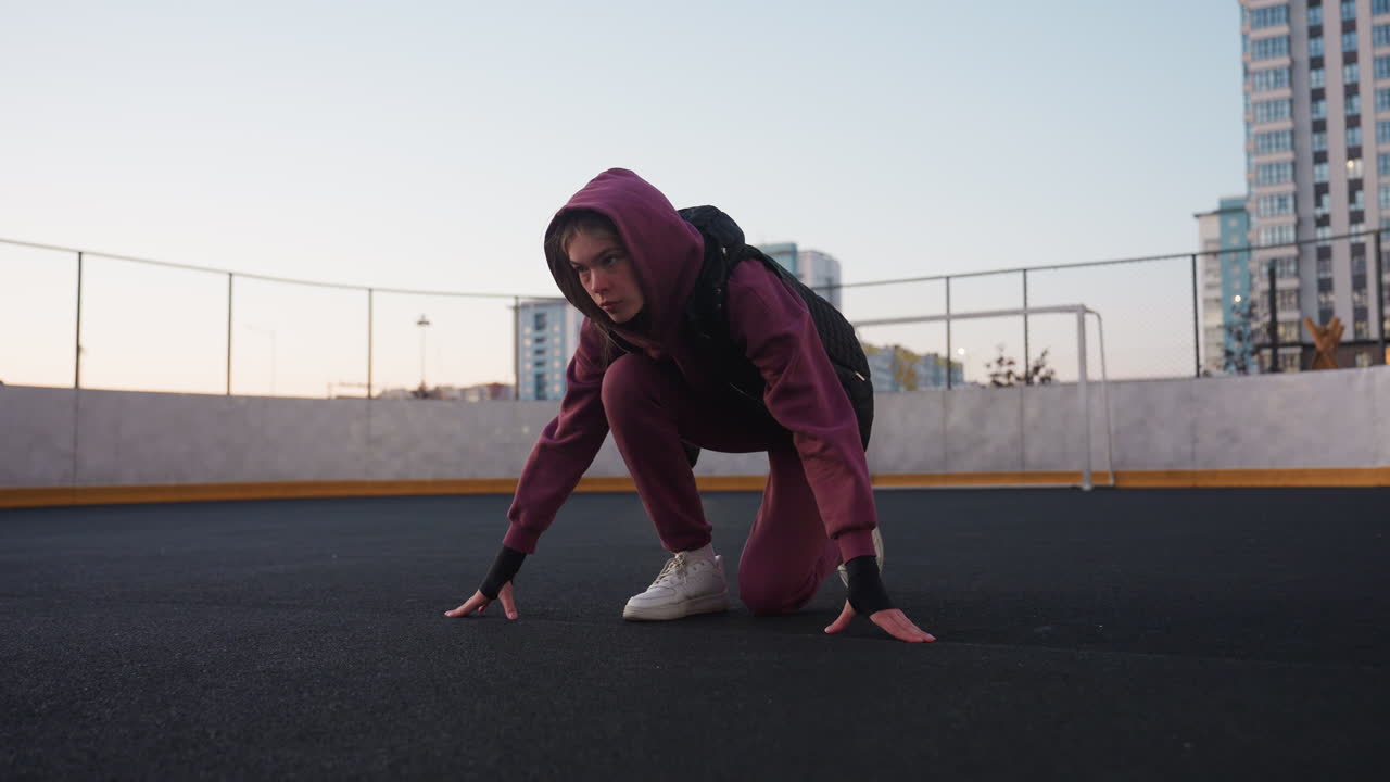 Active woman kneeling in starting position with hands on ground on curved sports court asphalt next to white barrier topped with chain link fence wearing maroon hoodie black vest and white sneakers