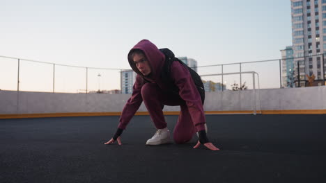 active woman kneeling in starting position with hands on ground on curved sports court asphalt next to white barrier topped with chain link fence wearing maroon hoodie black vest and white sneakers