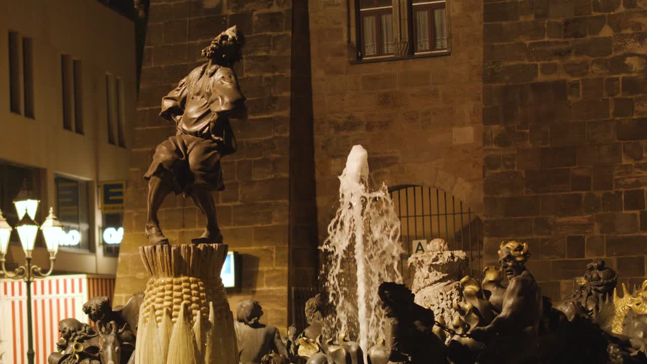 Illuminated fountain statue with corn motif at night, water spraying, warm lighting, steady camera