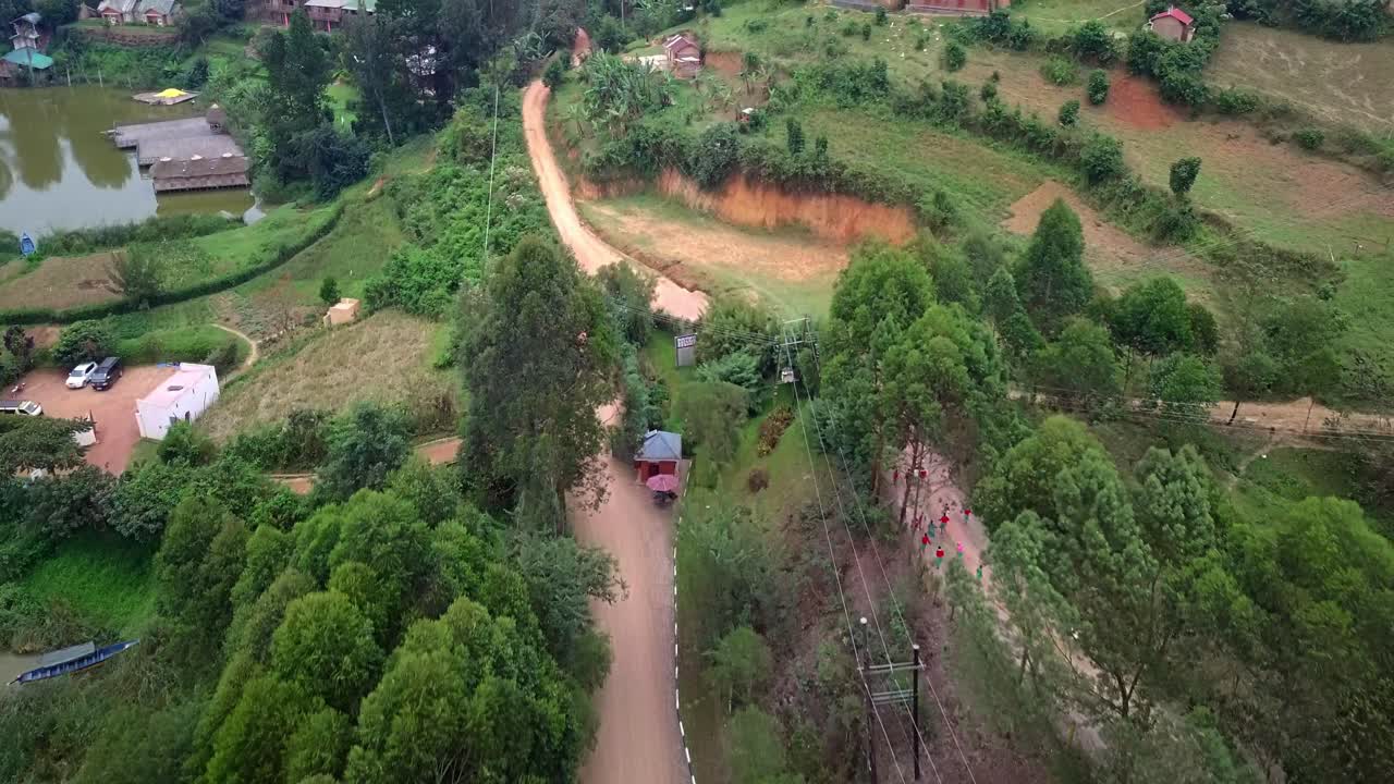 Top-down drone image of terraced agricultural slopes and scattered homes along Lake Bunyonyi’s shoreline in Uganda, showing winding dirt roads, small water inlet, lush vegetation and rural patterns