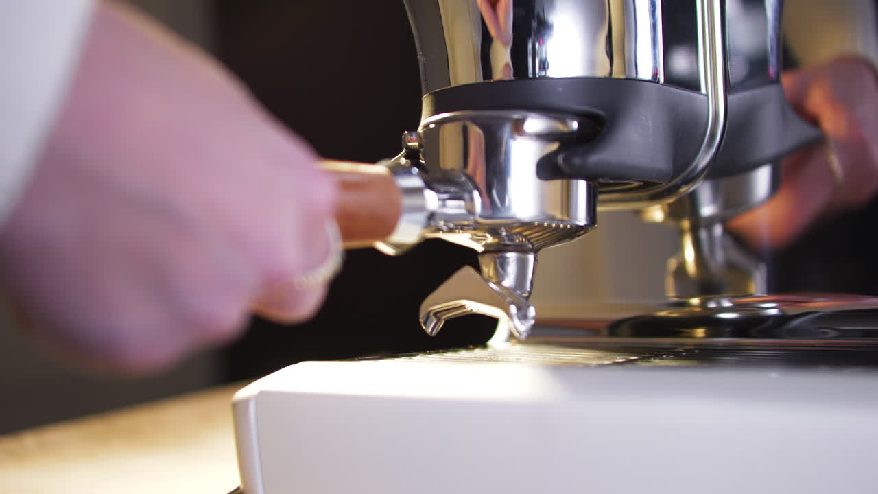 Primo piano di un barista che inserisce un portafiltro con manico in legno in un macinacaffè automatico per dosare il caffè macinato fresco. Movimenti precisi e riflessi metallici evidenziano il flusso di lavoro e la preparazione.