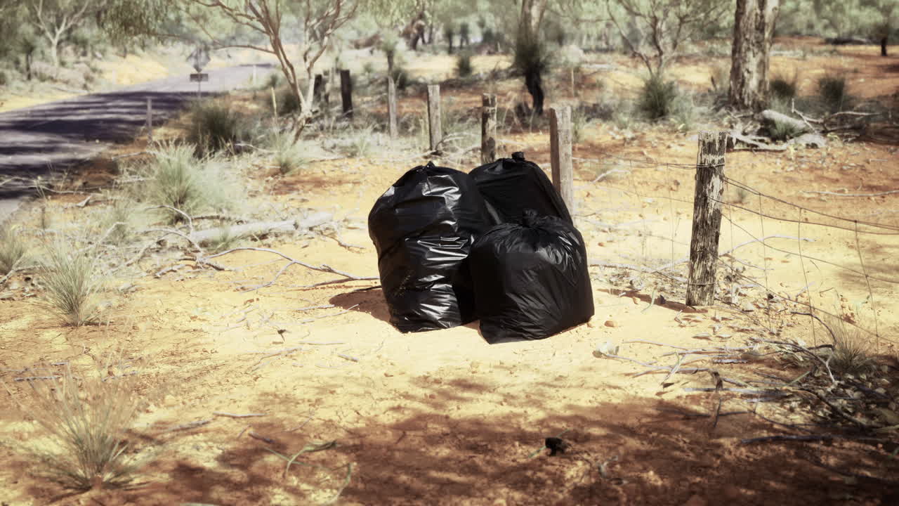 Trash bags left near a dirt road surrounded by dust and trees