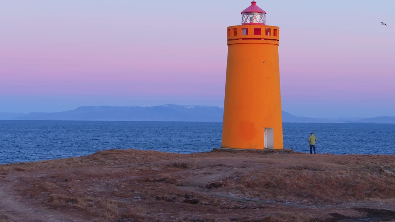 Un vivace faro arancione si staglia contro un cielo crepuscolare viola sulla costa frastagliata dell'Islanda, con una persona solitaria che guarda il mare