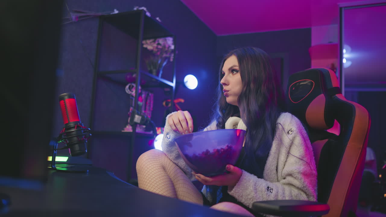 Female programmer sits in ergonomic gaming chair with crossed legs, eating popcorn from large bowl under colorful studio lights while watching screen in tech-inspired modern workspace
