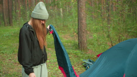 woman wearing gray beanie and black jacket walks toward tent holding peg while assisting husband during camping setup in lush forest with tall trees and natural greenery all around
