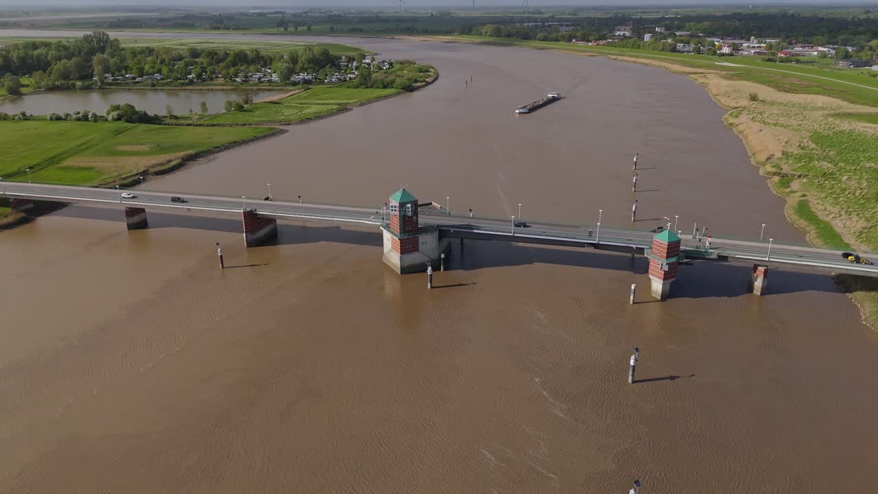 Drone footage showing a freight vessel cruising along the Ems River near the Jann-Berghaus Bridge in Leer, Germany. Captured from a steady frontal perspective with road traffic and riverscape.