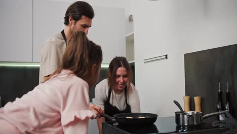 una chica morena feliz con un delantal negro prepara el desayuno con su novio y su hija pequeña por la mañana en una cocina moderna. una familia feliz preparando el desayunado por la mañanas en la cocina de un apartamento moderno