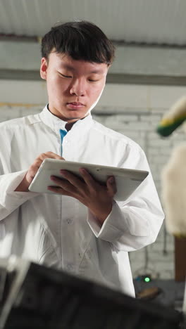 Mechanic demonstrates tool usage on engine while students take notes on tablets, second mechanic observes student&#039;s input, industrial automotive workshop filled with tools, machinery in background