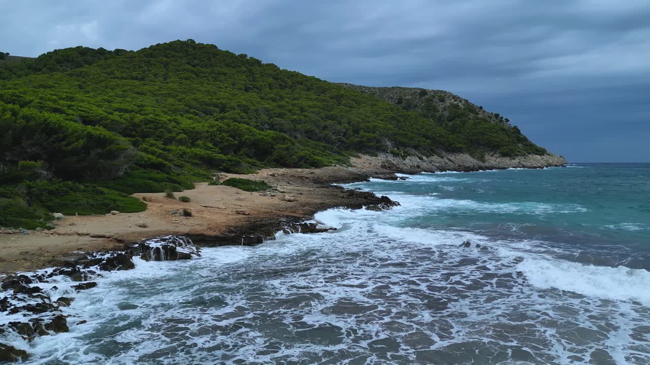 Dramatic high-angle drone shot capturing Cala Agulla beach and its rugged coves in Majorca. Heavy waves under a dark, cloudy sky, highlighting the powerful natural landscape